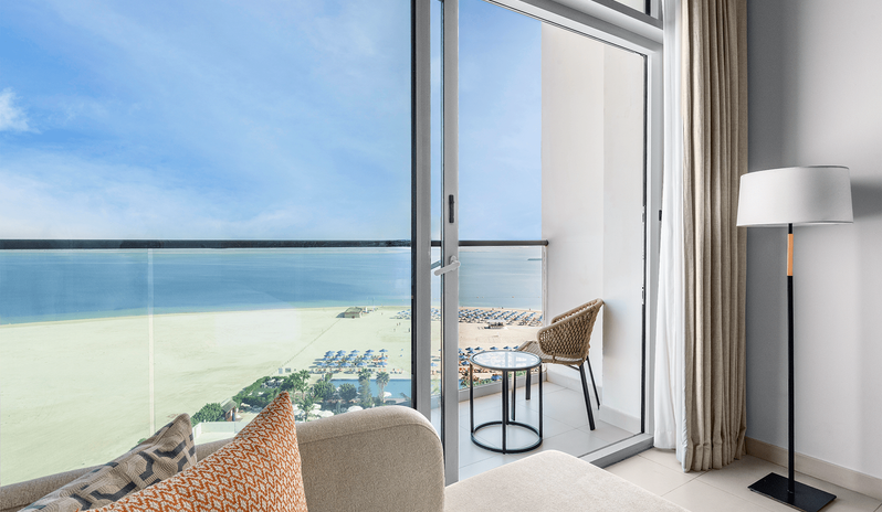 Family Room Panoramic Sea View Double Double sliding glass doors from a hotel suite seating area onto a private balcony with a small table, wicker chair, and beach view at Centara Mirage Beach Resort Dubai by Kenwood Travel