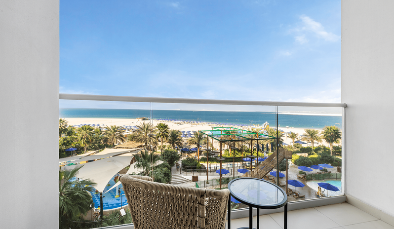 Family Room Sea View Double Double Private balcony with a woven chair and glass table, overlooking the beach, outdoor pools, and ocean water park at Centara Mirage Beach Resort Dubai by Kenwood Travel