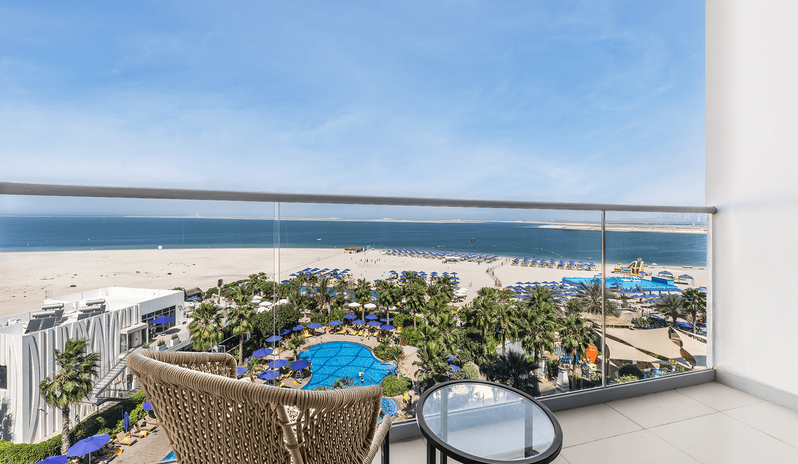 Mirage Family Room Panoramic Sea View with Bunk Beds Private hotel balcony with a woven chair and glass table, featuring an elevated view of a beachfront water park resort and the open sea at Centara Mirage Beach Resort Dubai by Kenwood Travel