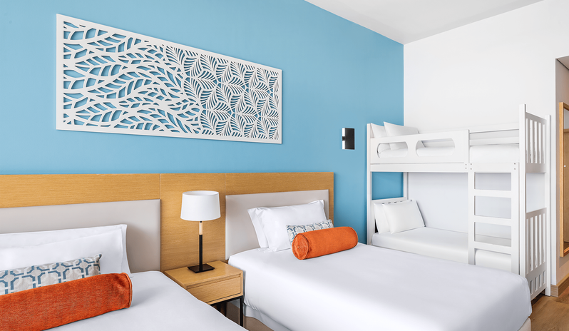 Mirage Family Room Sea View with Bunk Beds two comfortable beds with orange bolster pillows, white wooden bunk beds in the corner, and a distinctive white carved leaf-pattern art piece mounted on a bright blue accent wall at Centara Mirage Beach Resort Dubai by Kenwood Travel
