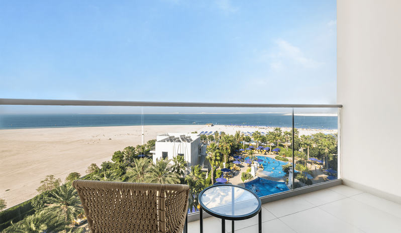 Mirage Family Room Sea View with Bunk Beds glass-panel railing, outdoor seating with a woven chair and glass-top table, overlooking the beach, outdoor pool complex, and the blue ocean at Centara Mirage Beach Resort Dubai by Kenwood Travel