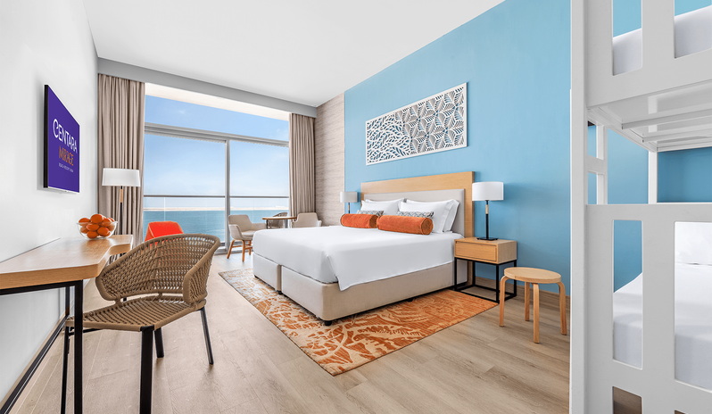 Mirage Superior Room Panoramic Sea View with Bunk Beds king bed with orange accent pillows, white bunk beds, a light blue accent wall with decorative artwork, a work desk with a woven chair, and large sliding glass doors opening to a panoramic sea view balcony at Centara Mirage Beach Resort Dubai by Kenwood Travel
