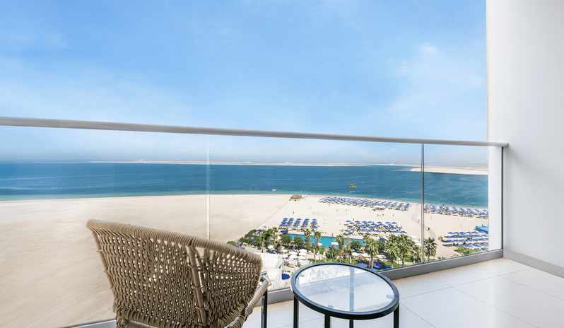 Mirage Superior Room Panoramic Sea View with Bunk Beds woven outdoor armchair and a glass-top side table against a glass railing, overlooking a wide sandy beach, a resort pool area with blue umbrellas, and a panoramic view of the blue sea and clear skyat Centara Mirage Beach Resort Dubai by Kenwood Travel