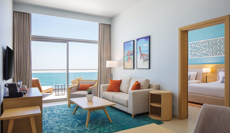 Luxury resort suite living area with a balcony overlooking the sea, a coffee table, and a glimpse into a connecting bedroom with a turquoise accent wall at Centara Mirage Beach Resort Dubai by Kenwood Travel