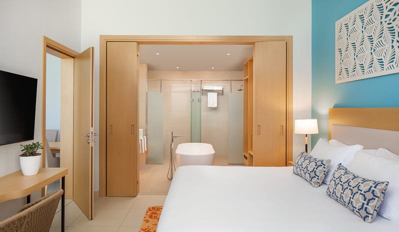Luxury resort suite bedroom featuring a white bed, teal accent wall, and a view into an open-concept bathroom with a modern soaking tub Luxury resort suite bedroom featuring a white bed, teal accent wall, and a view into an open-concept bathroom with a modern soaking tub at Centara Mirage Beach Resort Dubai by Kenwood Travel
