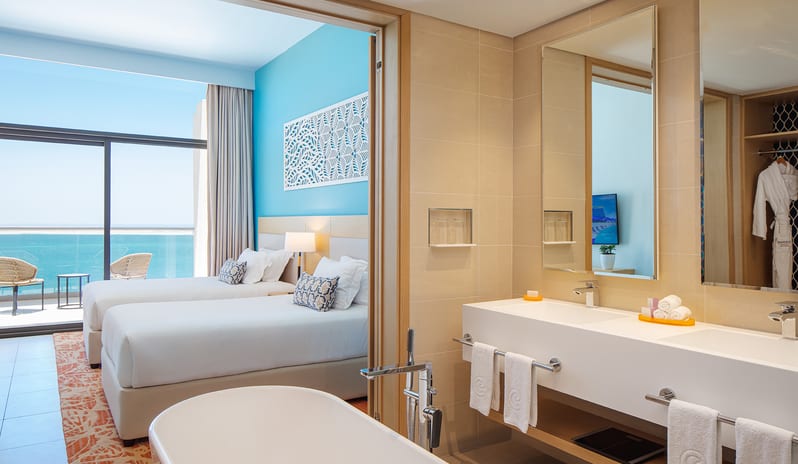 Luxury resort suite bathroom with a double sink vanity and bathtub, offering a view into an adjacent twin bedroom with a sea-facing balcony at Centara Mirage Beach Resort Dubai by Kenwood Travel