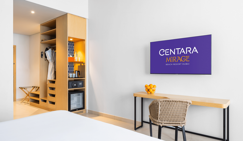 Superior Room Panoramic Sea View King room workspace and storage setup featuring a light-wood desk with a woven armchair, electronic in-room safe, mini bar, and hanging bathrobes at Centara Mirage Beach Resort Dubai by Kenwood Travel
