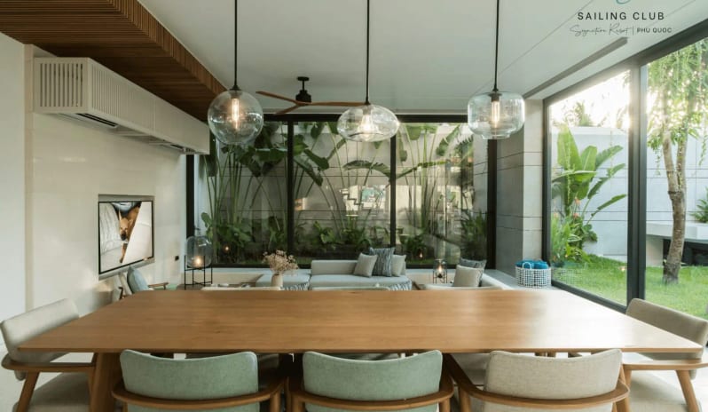 One Bedroom Pool Villa dining room interior with contemporary wooden chairs, minimalist decor, and a sunken living room area overlooking lush green foliage at Sailing Club Signature Resort Phu Quoc Hotel by Kenwood Travel