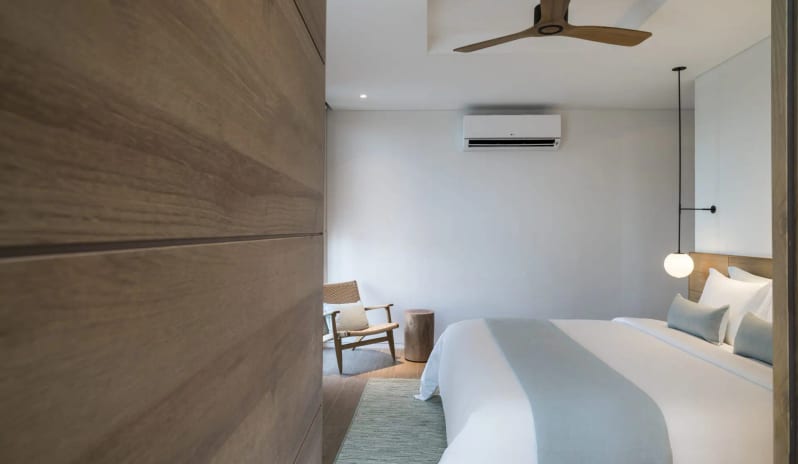 Signature Two Bedroom Deluxe Pool Villa bedroom featuring a wood-paneled entrance, a hanging globe bedside light, a modern air conditioner, and light wood furniture accents at Sailing Club Signature Resort Phu Quoc Hotel by Kenwood Travel