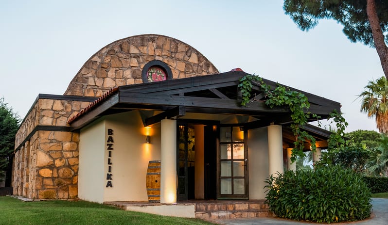 Bazilika A La Carte Restaurant Charming resort restaurant building with rustic stone masonry, a decorative wine barrel, and warm evening lighting at Gloria Verde by Kenwood Travel