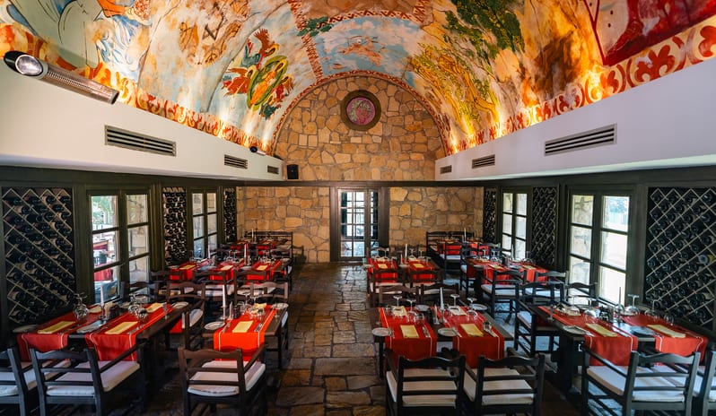 Bazilika A La Carte Restaurant Rustic resort wine cellar restaurant with flagstone flooring, built-in wine racks, set dining tables, and painted vaulted ceiling at Gloria Verde by Kenwood Travel