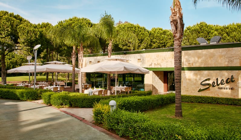 Select Restaurant featuring outdoor seating with large umbrellas, palm trees, and lush green hedges at Gloria Verde by Kenwood Travel