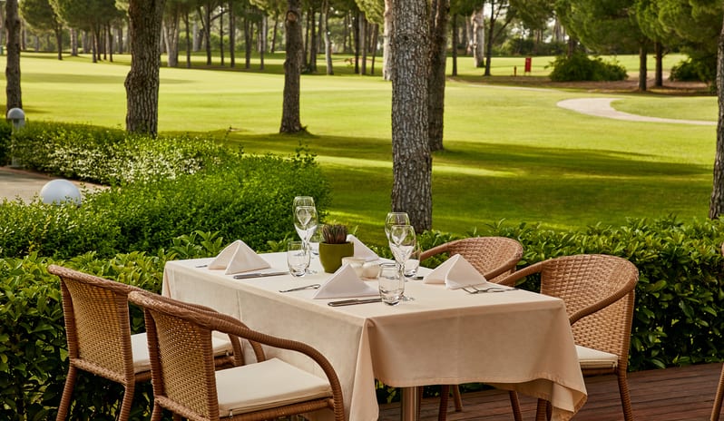 Select Restaurant table setting with white tablecloth, wine glasses, and folded napkins on a wooden deck next to a scenic golf course view at Gloria Verde by Kenwood Travel