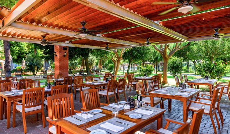 La Trattoria Romana A La Carte Restaurant wooden tables and chairs set for dinner under a large wooden pergola with ceiling fans, surrounded by a green garden at Gloria Serenity Resort by Kenwood Travel