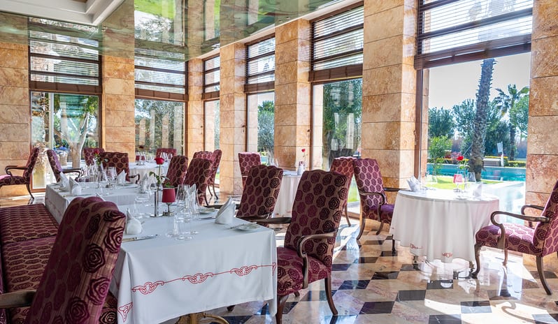 Le Jardin Au Printemps A La Carte Restaurant dining tables with white tablecloths, patterned maroon high-back chairs, a polished diamond-pattern floor, and a wooden service cart with wine at Gloria Serenity Resort by Kenwood Travel