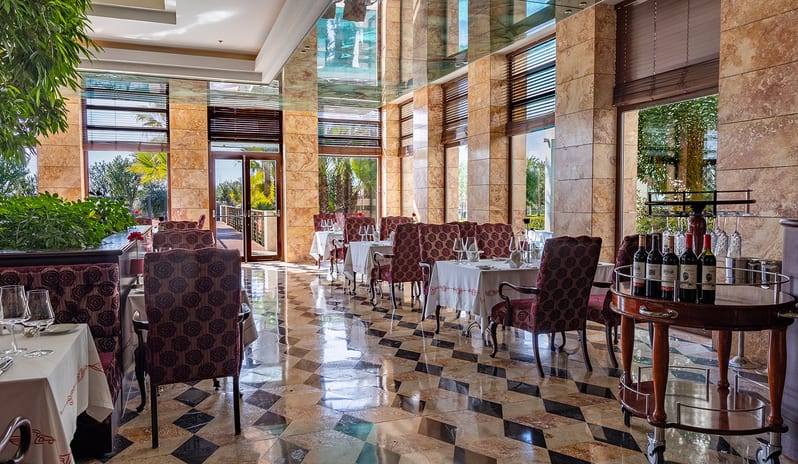 Le Jardin Au Printemps A La Carte Restaurant dining tables with white tablecloths, patterned maroon high-back chairs, a polished diamond-pattern floor, and a wooden service cart with wine at Gloria Serenity Resort by Kenwood Travel