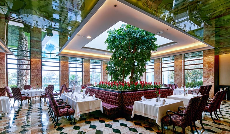 Le Jardin Au Printemps A La Carte Restaurant indoor garden atrium with a large central tree, red flowers, tufted velvet booths, white-clothed dining tables, and a glossy patterned floor at Gloria Serenity Resort by Kenwood Travel