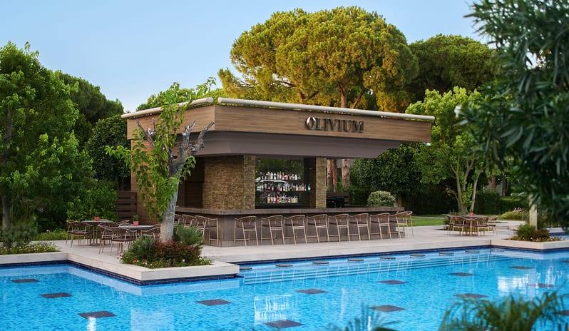Olivium Pool Bar modern wood and stone open-air counter with bar stools alongside a vibrant blue mosaic-tiled swimming pool surrounded by lush green trees at Gloria Serenity Resort by Kenwood Travel