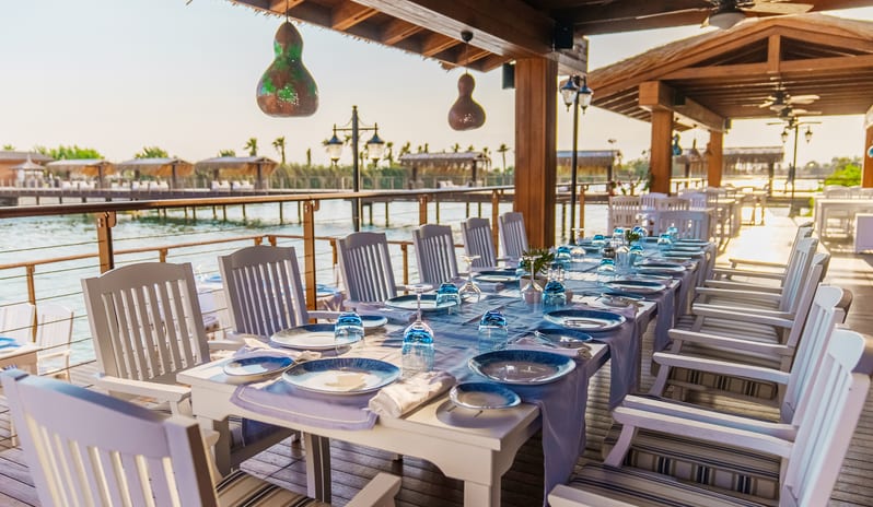 River Landing A La Carte Restaurant dinner with fine tableware, a long wooden table, striped seat cushions, and decorative lighting under a rustic wood pavilion over a river at Gloria Serenity Resort by Kenwood Travel