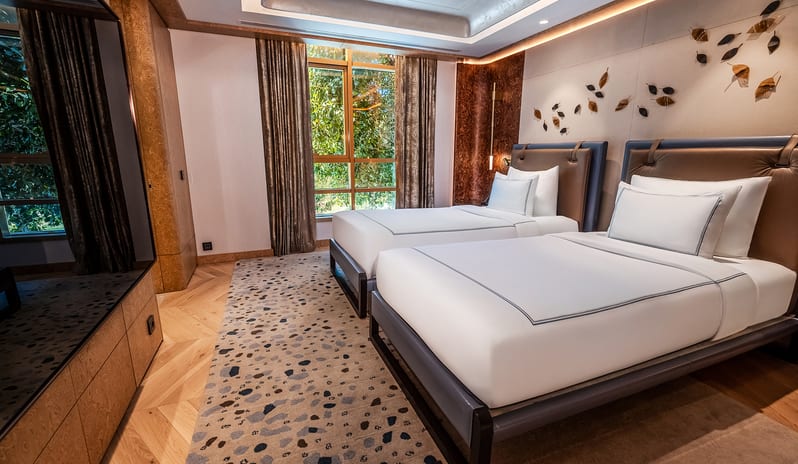 Vip Villa twin bedroom interior with light wood chevron floors, crisp white hotel bedding, built-in wooden wardrobes, and a large window looking out onto greenery at Gloria Serenity Resort by Kenwood Travel