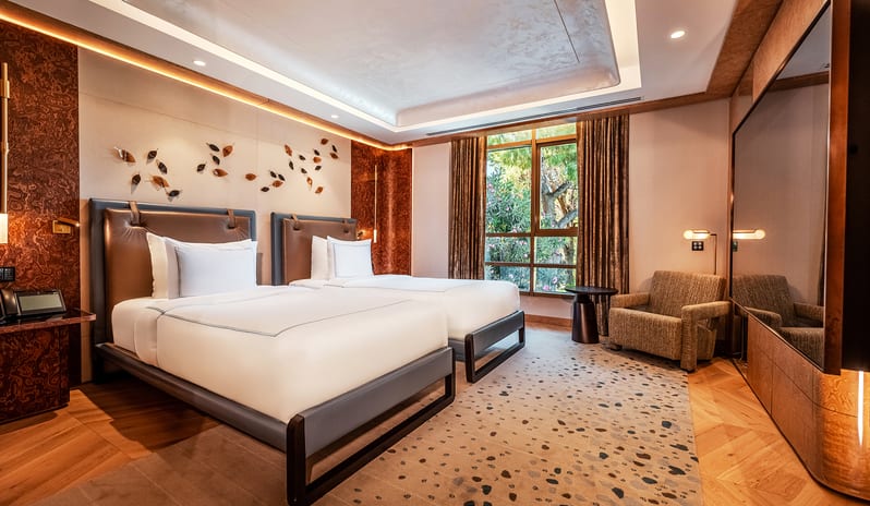 Vip Villa two single beds with brown leather headboards, a textured armchair, a large gloss black TV media wall, and a window with garden views at Gloria Serenity Resort by Kenwood Travel