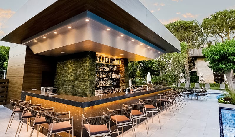 Olivium Pool Bar stone-clad pillar, fully stocked back bar shelves, under-counter ambient lighting, woven high-back bar stools, and a poolside deck at dusk at Gloria Serenity Resort by Kenwood Travel