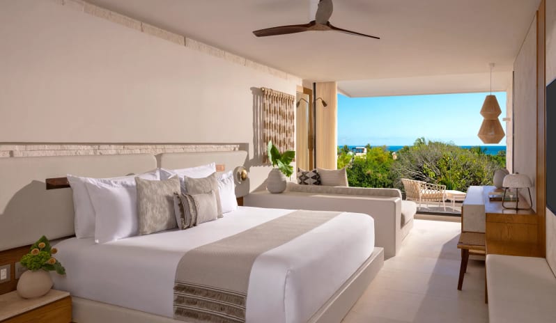 Junior Suite King Partial Ocean View neutral boho decor, a plush bed with textured pillows, a ceiling fan, a private indoor daybed sofa, and a wide open balcony looking out over lush green trees and the blue ocean at Impression Isla Mujeres by Secrets by Kenwood Travel