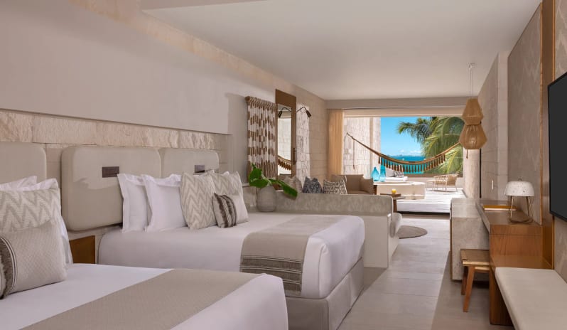 Signature Junior Suite Double Ocean View W/ Hot Tu luxury hotel room with white stone walls, natural wood furniture, a built-in daybed sofa, and a private balcony with outdoor seating overlooking a tropical beach landscape at Impression Isla Mujeres by Secrets by Kenwood Travel