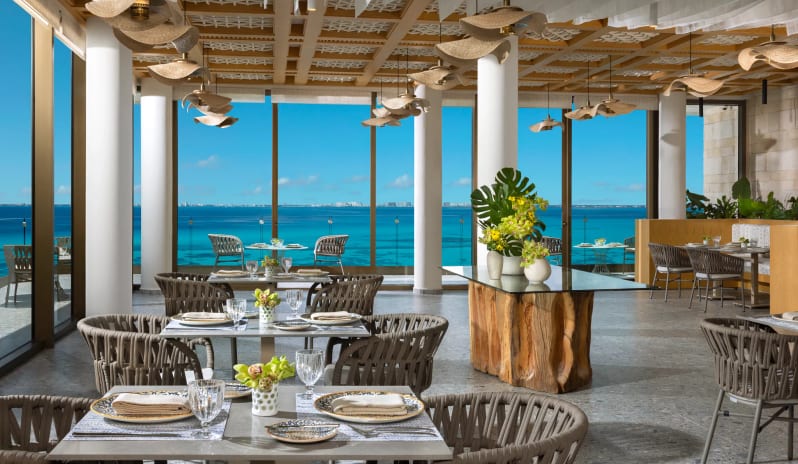 La Vista contemporary coastal interior design, a central live-edge wood table with tropical floral arrangements, and panoramic sea view at Impression Isla Mujeres by Secrets by Kenwood Travel