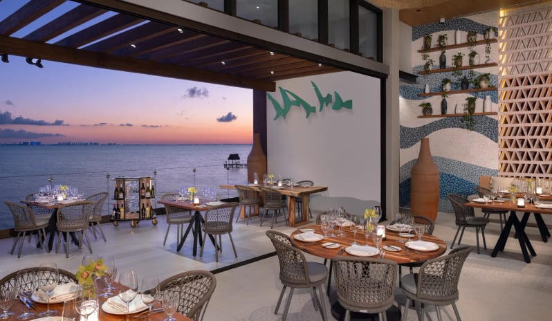 Spezia open-air dining area with round wooden tables, contemporary woven chairs, and a stunning sunset view over the ocean at Impression Isla Mujeres by Secrets by Kenwood Travel