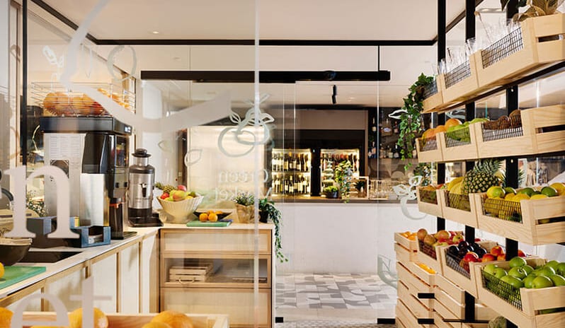 Quattro Terre Wooden shelves lined with crates of fresh apples, oranges, and pineapples next to a coffee and juice machine inside the modern Green Market shop at Pical Resort, Valamar Collection by Kenwood Travel