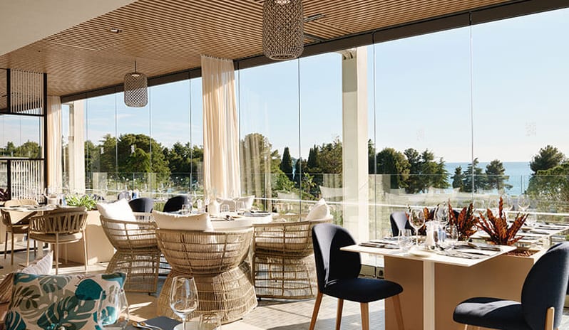 More woven armchairs, large glass windows, and scenic views of the surrounding trees and sea at Pical Resort, Valamar Collection by Kenwood Travel