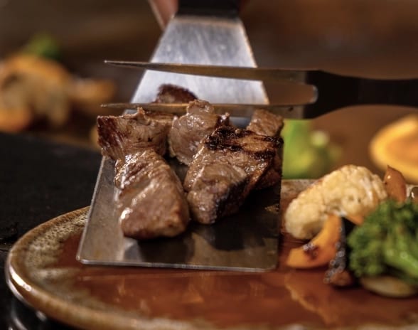 Teppan by Momo Teppanyaki chef serving grilled steak cubes with a metal spatula onto a plate at Moon Palace The Grand Punta Cana by Kenwood Travel