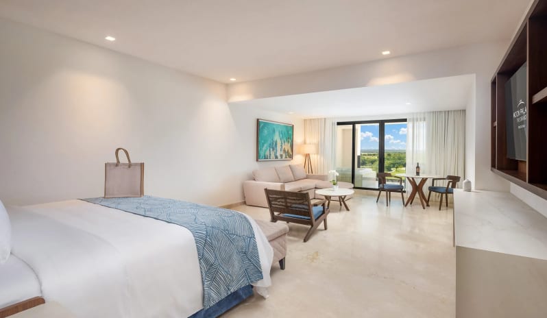Grand Junior Suite Golf View king bed with a blue pattern runner, an adjoining living area with a sofa, armchair, dining set, and sliding doors leading to a golf view balcony at Moon Palace The Grand Punta Cana by Kenwood Travel