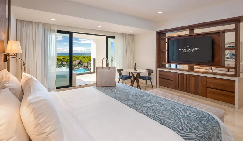 Grand Governor Suite Ocean View king-size bed, wooden TV console partition, dining table for two, and open balcony sliding doors showing a tropical ocean view at Moon Palace The Grand Punta Cana by Kenwood Travel