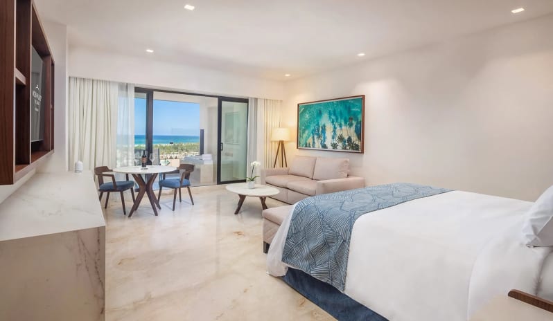 Grand Family Suite Ocean View sofa lounge area, dining table for two, and sliding glass balcony doors looking out to the ocean at Moon Palace The Grand Punta Cana by Kenwood Travel