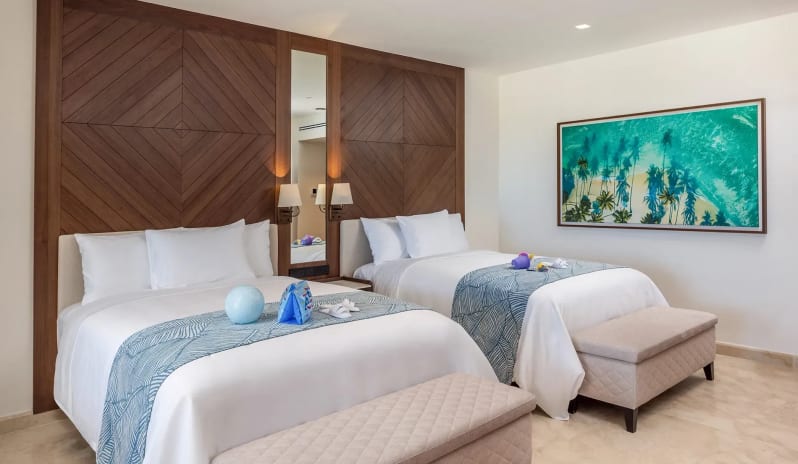 Grand Family Suite Golf View bedroom with two double beds against a dark wood accent wall, upholstered end-of-bed storage benches, and bright turquoise beach artwork on the wall at Moon Palace The Grand Punta Cana by Kenwood Travel