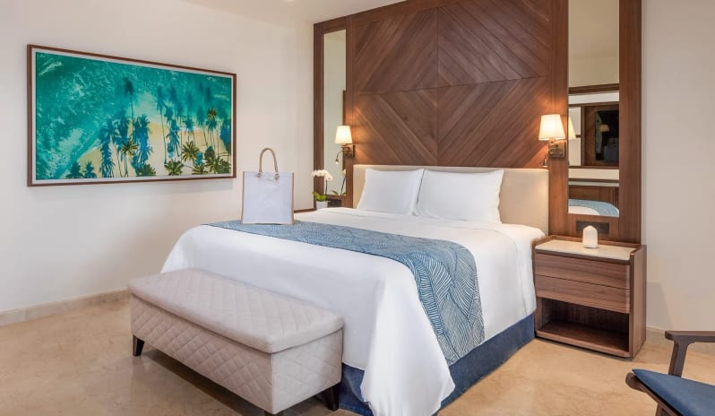 Grand Governor Suite Ocean View king-size bed dressed in white linens, an upholstered storage bench at the foot, warm bedside lamps, and a large tropical beach art print on the wall at Moon Palace The Grand Punta Cana by Kenwood Travel