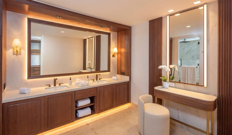 Grand Governor Suite Ocean View bathroom with dark wood double sinks, under-cabinet ambient lighting, a large backlit mirror, and a separate vanity makeup console next to fluted wall panels at Moon Palace The Grand Punta Cana by Kenwood Travel
