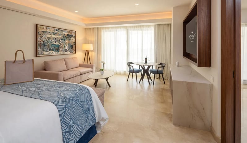Grand Presidential Suite Golf View bedroom interior showing a corner view of a white-draped bed, cozy sofa area with framed artwork above, a small two-seat dining table, and light marble flooring at Moon Palace The Grand Punta Cana by Kenwood Travel