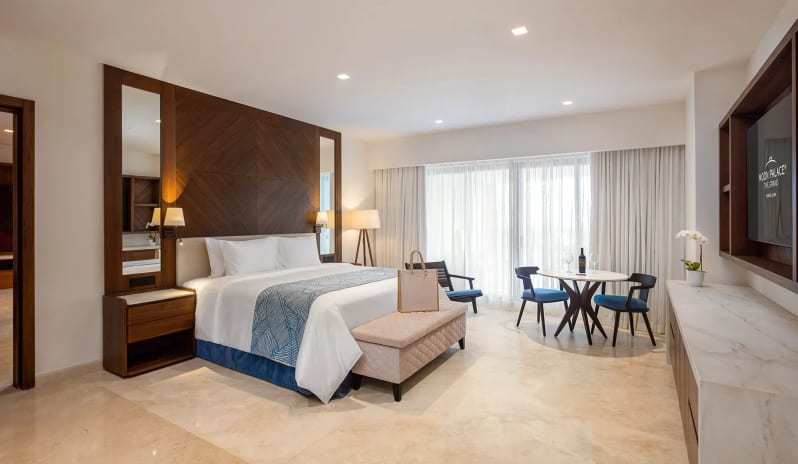 Grand Presidential Suite Golf View master bedroom with a king bed dressed in crisp white linens, a padded ottoman bench, small bistro table with two chairs, and sheer curtains covering floor-to-ceiling balcony glass doors at Moon Palace The Grand Punta Cana by Kenwood Travel