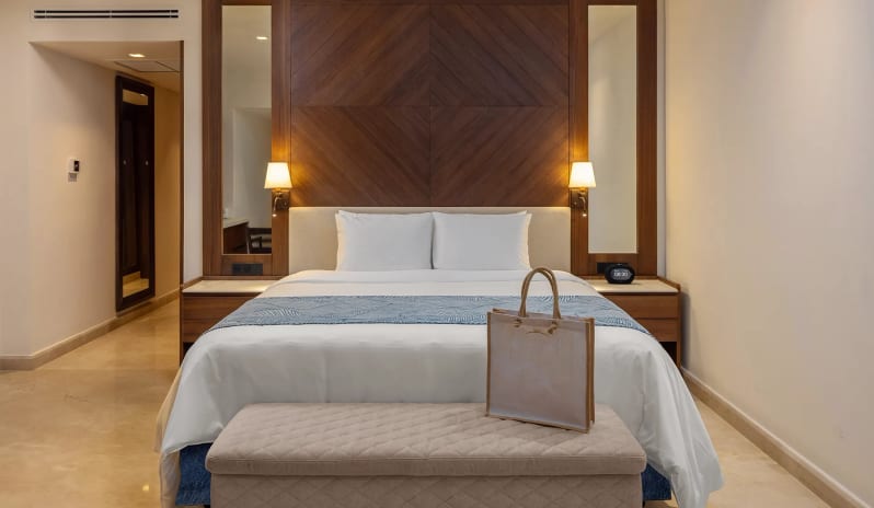 Grand Swim-up Suite king-size bed with a wooden accent headboard, bedside sconces, and a tote bag on a bench at Moon Palace The Grand Punta Cana by Kenwood Travel