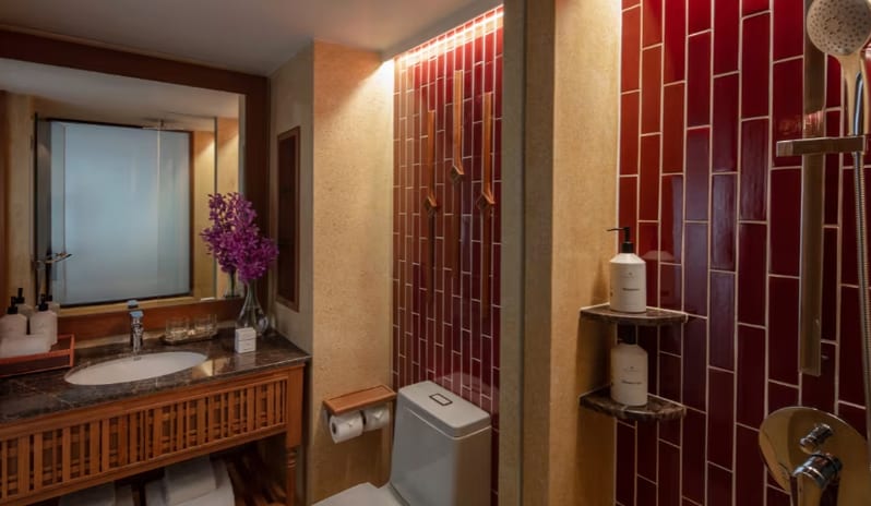 Deluxe Pool Access bathroom featuring a wooden vanity with marble top, large mirror, purple orchid accents, toilet, and a walk-in shower area lined with glossy vertical red tiles at Anantara Hua Hin Resort  by Kenwood Travel