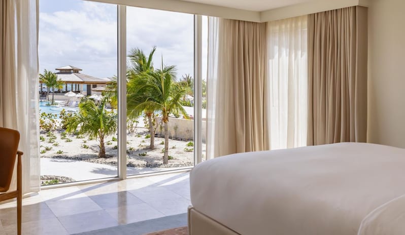 Two Bedroom Ocean View Suite Lanai Patio King bedroom in a resort suite with large floor-to-ceiling windows overlooking palm trees and pool grounds at Salterra, A Luxury Collection Resort & Spa, South Caicos by Kenwood Travel