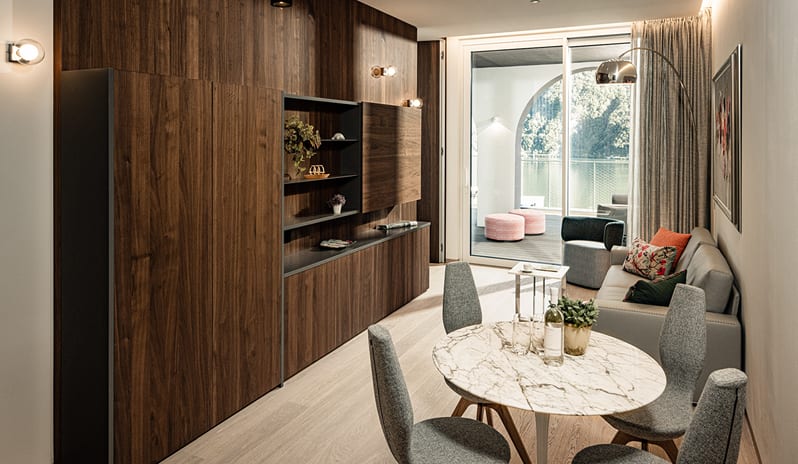 Lake View One Bedroom Loggia Suite living space with a round marble top dining table, grey upholstered chairs, rich dark wood storage cabinets, and floor-to-ceiling glass doors opening to a scenic lake terrace at & SPA by Kenwood Travel
