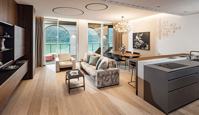 Lake View Grand Loggia Suiteliving area with a dark kitchen island, upholstered sofa, dining setup, and large sliding glass doors leading to a private loggia balcony with lake views at ARIA Retreat & SPA by Kenwood Travel