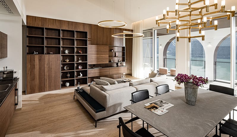 Lake View Family Grand Loggia Suite living room with a neutral L-shaped sofa, dark wood kitchen cabinetry, marble top coffee table, oval dining table, and glass doors to a private terrace at ARIA Retreat & SPA by Kenwood Travel