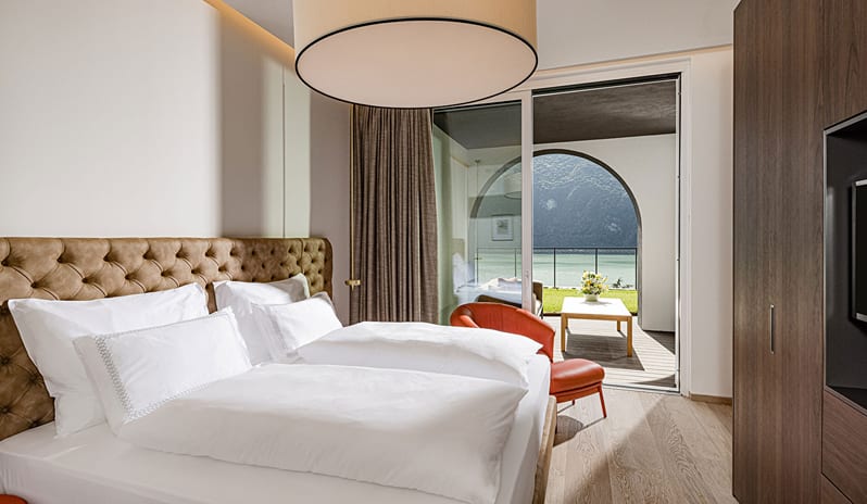 Lake View Family Grand Loggia Suite bedroom with crisp white bedding, tufted headboard, red leather lounge chair, and sliding glass doors leading to a private lake-view terrace with arched architecture at ARIA Retreat & SPA by Kenwood Travel