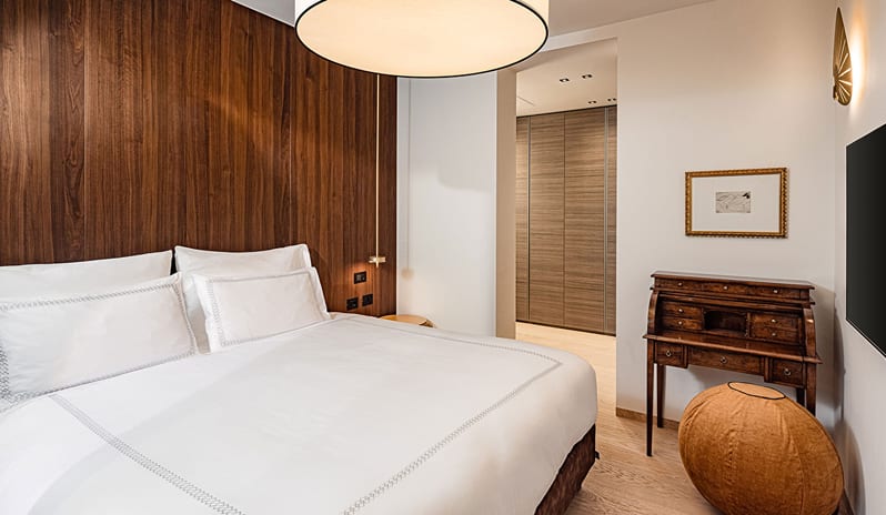 Lake View Grand Loggia Suite bedroom with crisp white bedding, rich wood wall paneling, antique-style secretary desk, leather pouf, and wall-mounted TV at ARIA Retreat & SPA by Kenwood Travel
