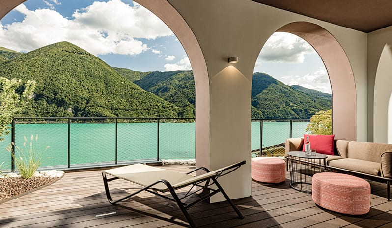 Lake View One Bedroom Loggia Suite private hotel suite balcony terrace framed by elegant wall arches, featuring outdoor lounge seating, sunbed, and panoramic views of green mountains across turquoise water at & SPA by Kenwood Travel